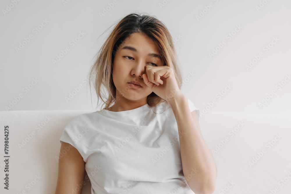 Sleepy face Asian woman yawning and rubbing her eyes in white t-shirt sits on the sofa. Sleepy face Asian woman yawning and rubbing her eyes in white t-shirt sits on the sofa.