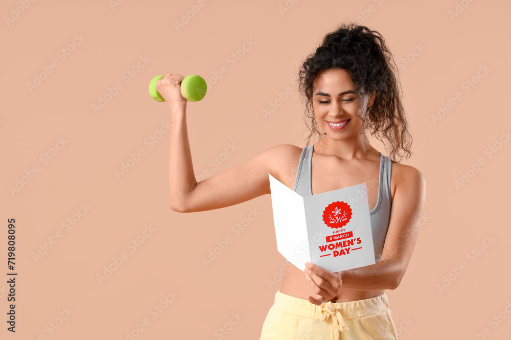 Young African-American woman in sportswear reading greeting card while exercising with dumbbell on beige background. International Women's Day celebration Young African-American woman in sportswear reading greeting card while exercising with dumbbell on beige background. International Women's Day celebration