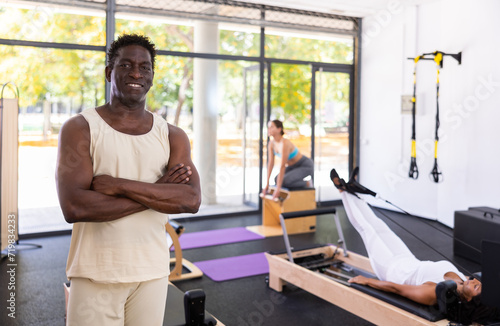 Positive African American man posing in fitness studio, clients perform exercise on background