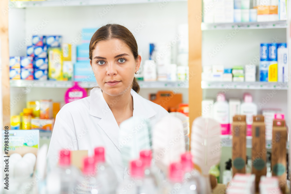 Female pharmacist in gown standing behind counter in drugstore. Shelves with medicine behind her.