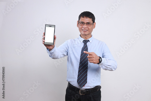 Cheerful adult Asian businessman in glasses and formal clothes pointing at smartphone with blank screen for mockup