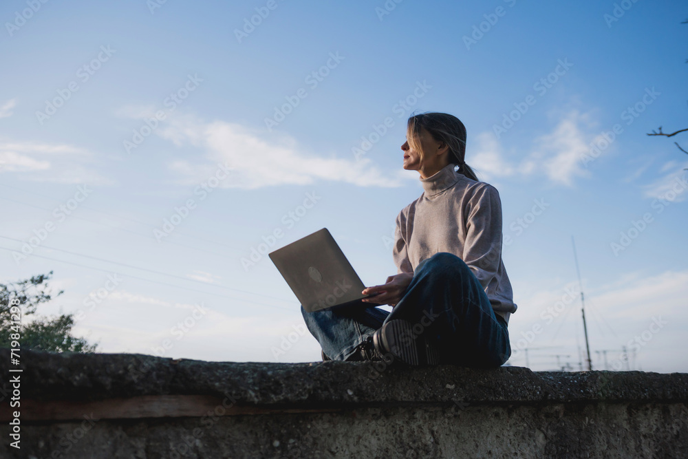 Woman freelancer uses laptop on cement wall outdoors against the sky. The woman to be focused on her work or enjoying some leisure time while using her laptop.