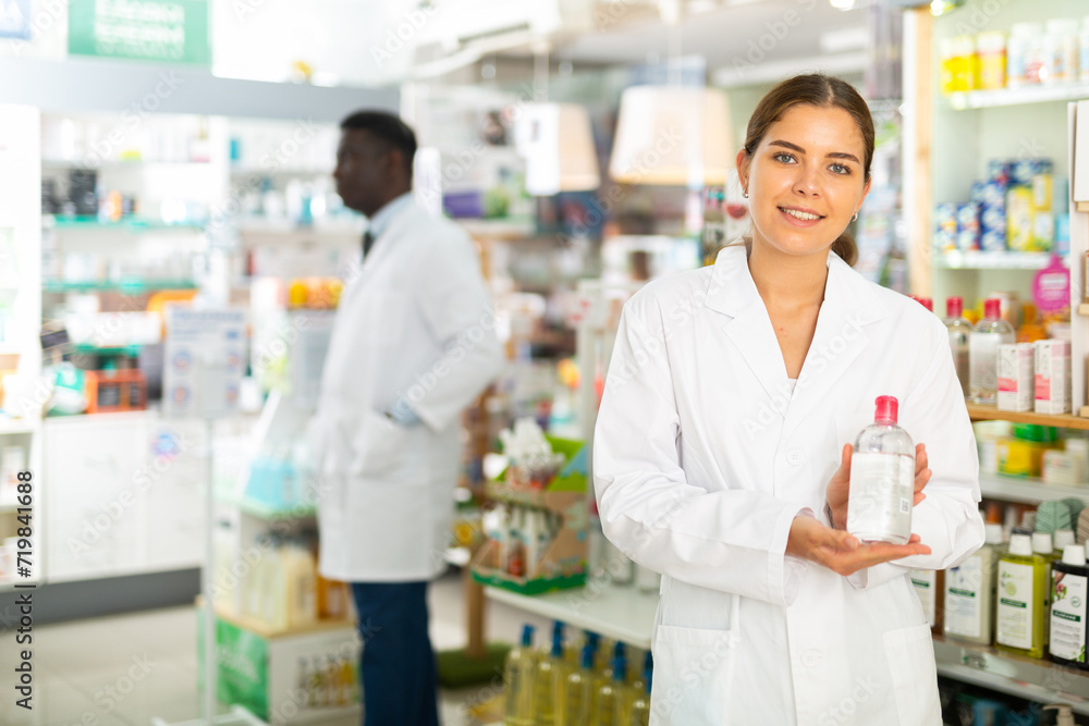 Female pharmacist in gown demonstrating skin care product in salesroom of drugstore