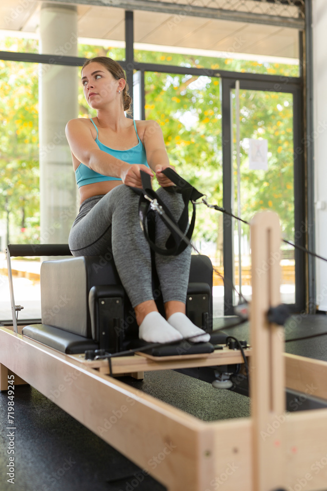 Fototapeta premium Girl stretching on reformer in gym. Caucasian young woman training pilates, using equipment.