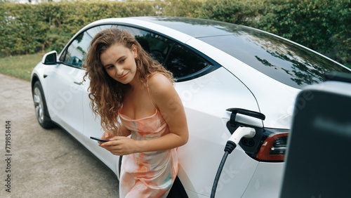 Young woman use smartphone to pay for electricity at public EV car charging station in nature. Modern environmental and sustainable automobile transportation lifestyle with EV vehicle. Synchronos © Summit Art Creations