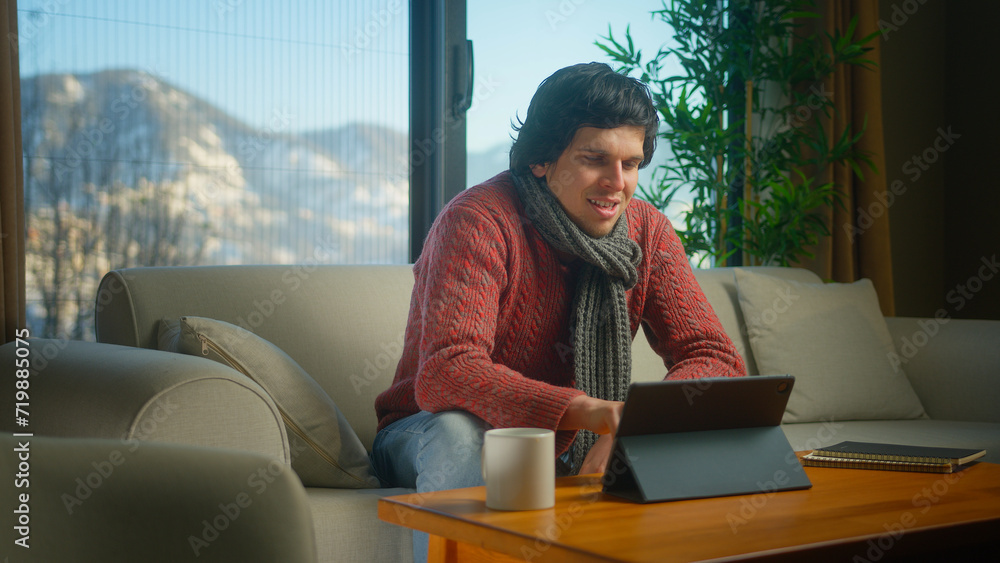 Smiling young man with scarf enjoying surfing internet, social media or watching video with tablet computer while sitting on sofa in living room. Modern tech device at home