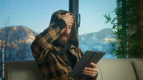 Young bearded man wear hoodie reacting to loss, bad news while using tablet computer. Man gets upset, feels anxiety and stress sitting on sofa in living room