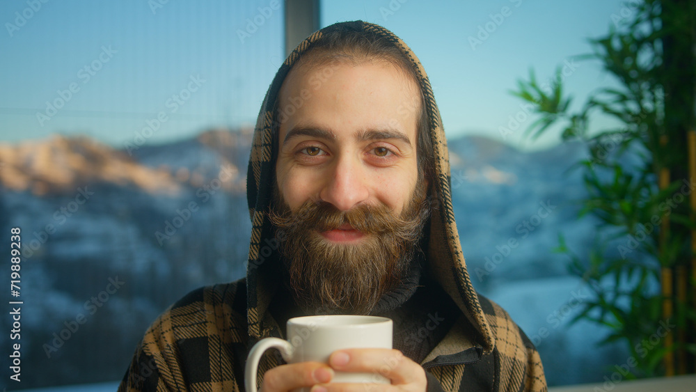 Portrait of bearded young man wearing hoodie drinking hot coffee or tea looking at camera smiling while sitting on couch in living room Portrait of bearded young man wearing hoodie drinking hot coffee or tea looking at camera smiling while sitting on couch in living room