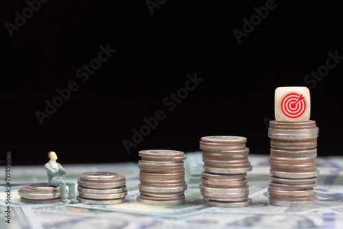 Pile of coins on banknote with figures models and target icon on wooden cube above stack of coins against dark background. Success business and financial growth by investment concept.