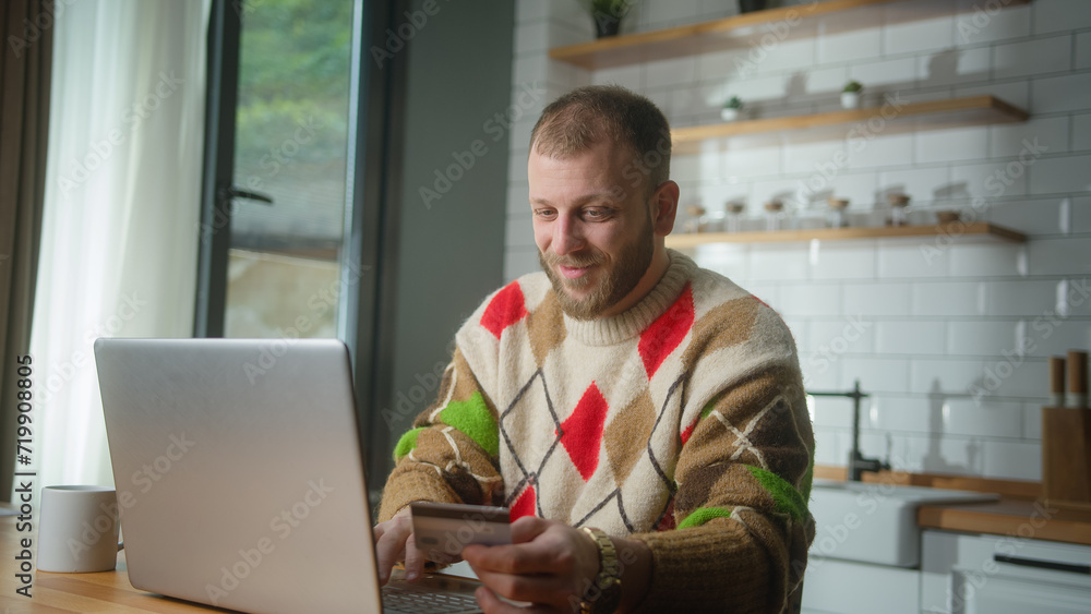 Smiling man enter credit card number on laptop for shopping online, makes secure easy distant electronic payment, gets happy while sitting in kitchen at home Smiling man enter credit card number on laptop for shopping online, makes secure easy distant electronic payment, gets happy while sitting in kitchen at home