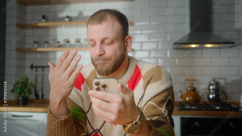 Man sitting in kitchen reacts to what sees, surfing internet on mobile phone, checking email, reading media news, scrolling social medias, using mobile applications on smartphone at home Man sitting in kitchen reacts to what sees, surfing internet on mobile phone, checking email, reading media news, scrolling social medias, using mobile applications on smartphone at home