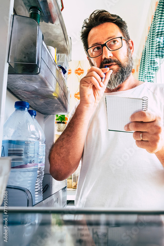 Adult caucasian man taking note list of food looking inside the open fridge at home - kitchen activity and alternative point of view - market shopping and quarantine lockdown stayhome coronavirus time photo