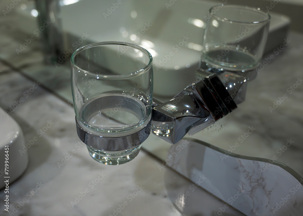 Transparent glass cup in a metal holder in the bathroom. In the background is a mirror with a reflection of a glass and a white sink.