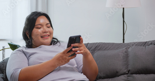 A chubby Asian woman using the mobile phone while relaxing on the sofa at home.