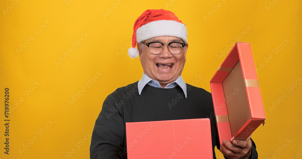 An old Asian man wearing a Santa hat and holding a large gift box, isolated on yellow background in the studio. An old Asian man wearing a Santa hat and holding a large gift box, isolated on yellow background in the studio.