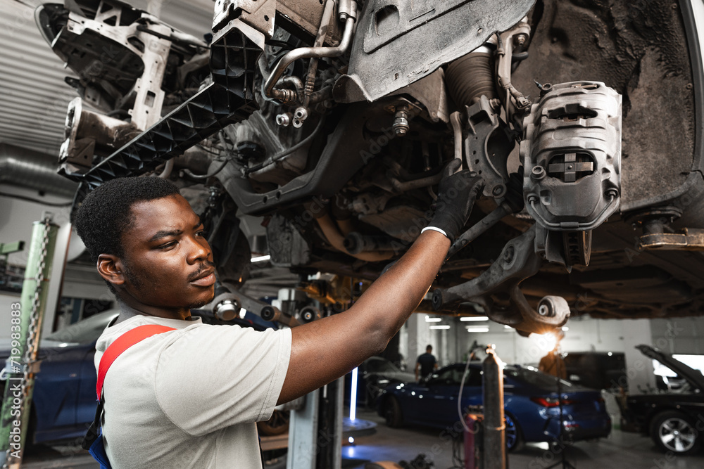 Young African mechanic in uniform working under the car in car service center Young African mechanic in uniform working under the car in car service center
