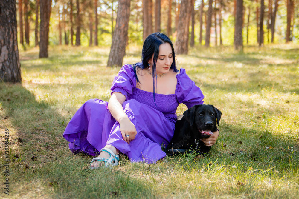 A young woman in a purple dress sits next to a black Labrador retriever against the backdrop of the park. A young woman in a purple dress sits next to a black Labrador retriever against the backdrop of the park.