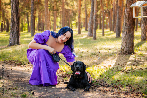 A young woman in a purple dress sits next to a black Labrador retriever against the backdrop of the park.