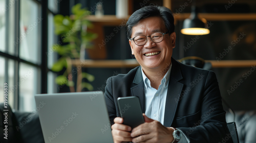 Senior middle aged Asian businessman manager ceo using smartphone and laptop in office working online Senior middle aged Asian businessman manager ceo using smartphone and laptop in office working online