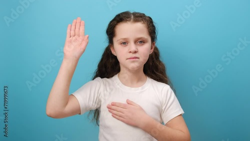 Portrait of serious preteen girl child raising her palm to take oath, child swearing to tell only truth, wearing white t-shirt, posing isolated over plain blue color background wall in studio photo