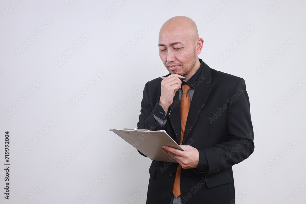 Young adult Asian bald man, business owner in suit looking at clipboard analyzing business report Young adult Asian bald man, business owner in suit looking at clipboard analyzing business report
