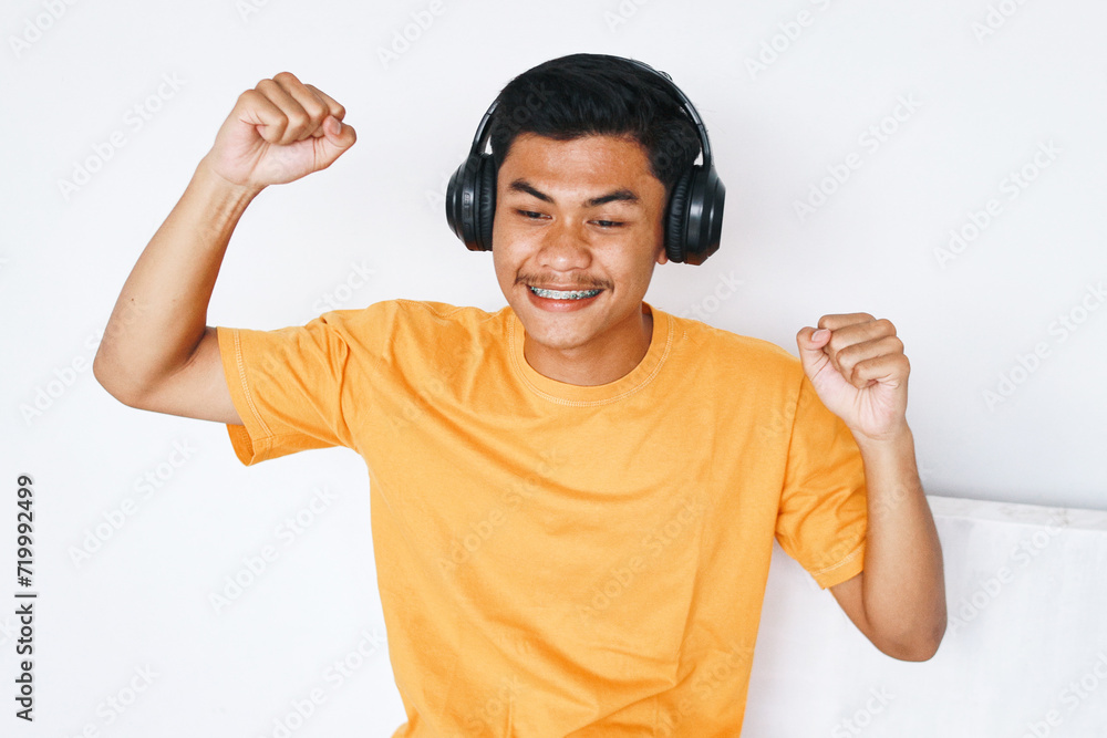 Fototapeta premium Happy Asian male student with braces teeth raising hands dancing while listening music from headphones
