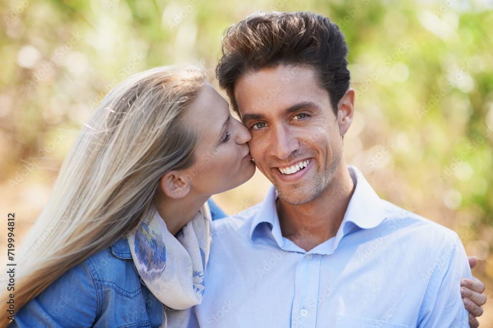 Happy couple, portrait and kiss on cheek in nature for love, bonding or support in relax for outdoor affection. Face of young woman and man smile for embrace, intimacy or romance in forest together