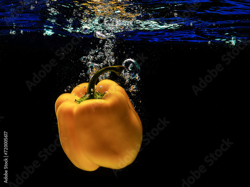 Yellow pepper falls into water with splashes and bubbles on a black background through the blue surface of the water.