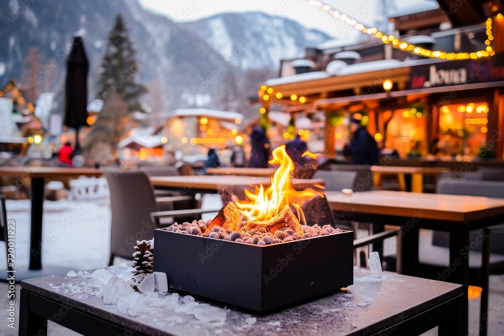 A cozy fire pit with a warm flame in a snowy outdoor setting at dusk, surrounded by festive lights and mountains. - obrazy, fototapety, plakaty 