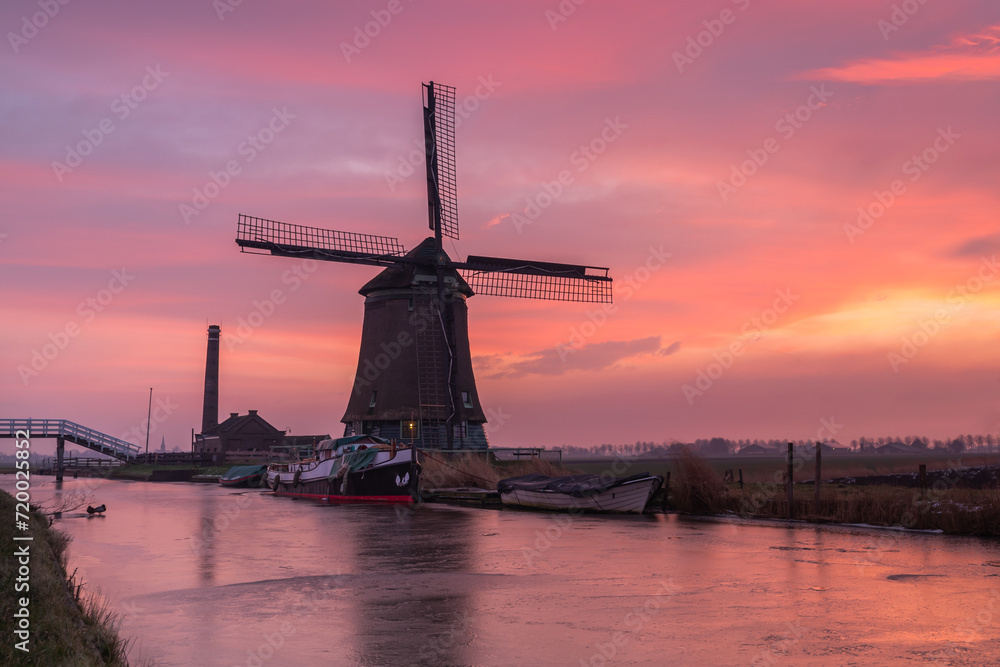 The Kaagmolen in Spanbroek (North Holland) under a brightly colored sky at sunrise. Boats line the canal banks, surrounded by a thin layer of ice after a moderate overnight frost.