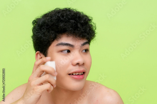 A cheerful young man with curly hair smiles while rubbing a bar of solid deodorant soap on his cheek. Facial care concept. Against a vivid green backdrop.