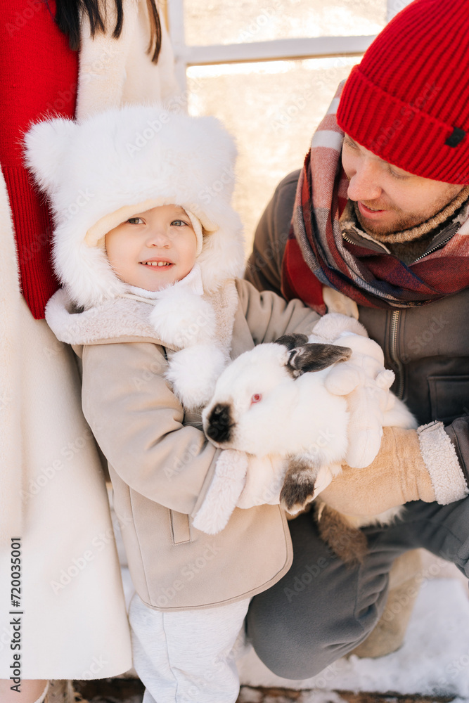 Obraz premium Vertical portrait of happy adorable son with loving family in warm clothes hugging white funny rabbit standing by house on sunny winter day. Concept of excursion to eco-farm, life in village.