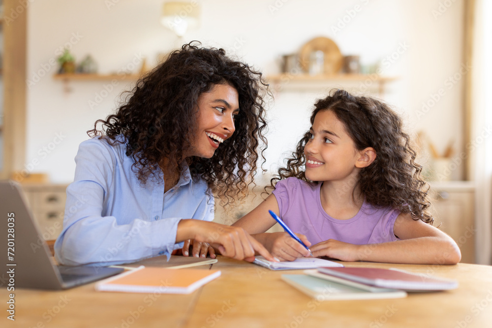 Education with mom concept. Mother helping daughter with homework, sitting in kitchen interior, at table with books and laptop Education with mom concept. Mother helping daughter with homework, sitting in kitchen interior, at table with books and laptop