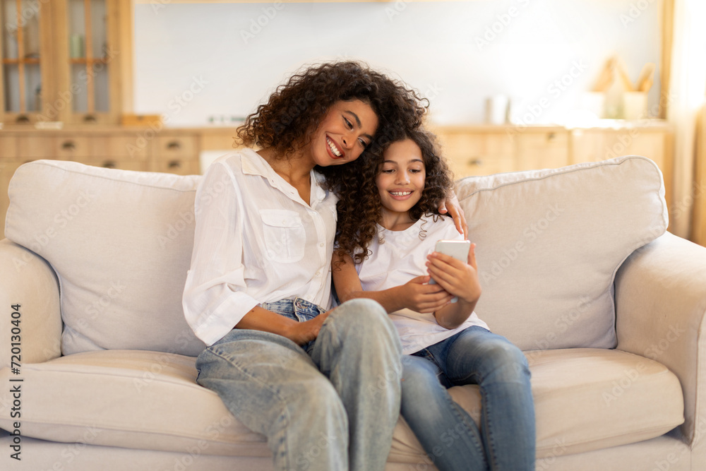Happy mother and little daughter using smartphone sitting on sofa at home, scrolling social media, websurfing via phone gadget Happy mother and little daughter using smartphone sitting on sofa at home, scrolling social media, websurfing via phone gadget