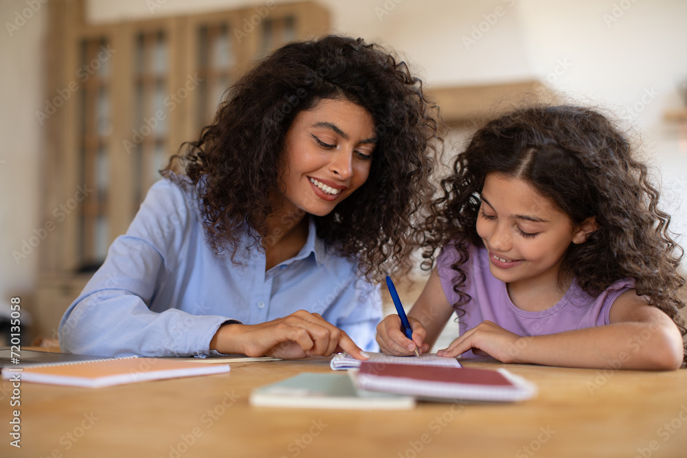 Caring young mom and her daughter sitting at table at home and making homework assignment together, tutor and girl child study learn do home task Caring young mom and her daughter sitting at table at home and making homework assignment together, tutor and girl child study learn do home task