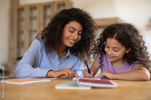 Caring young mom and her daughter sitting at table at home and making homework assignment together, tutor and girl child study learn do home task photo