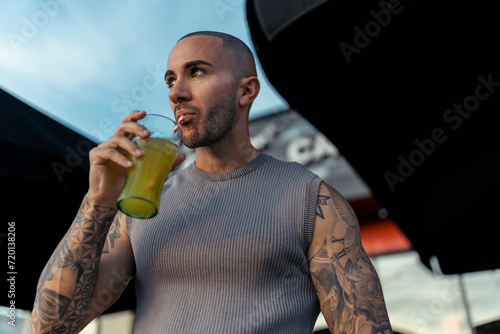 Chico joven musculado y tatuado posando con ropa urbana en terraza de restaurante de comida rapida bebiendo un refresco de naranja 
