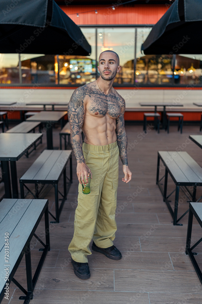 Chico tatuado y musculoso posando sin camiseta en terraza de restaurante de comida rapida