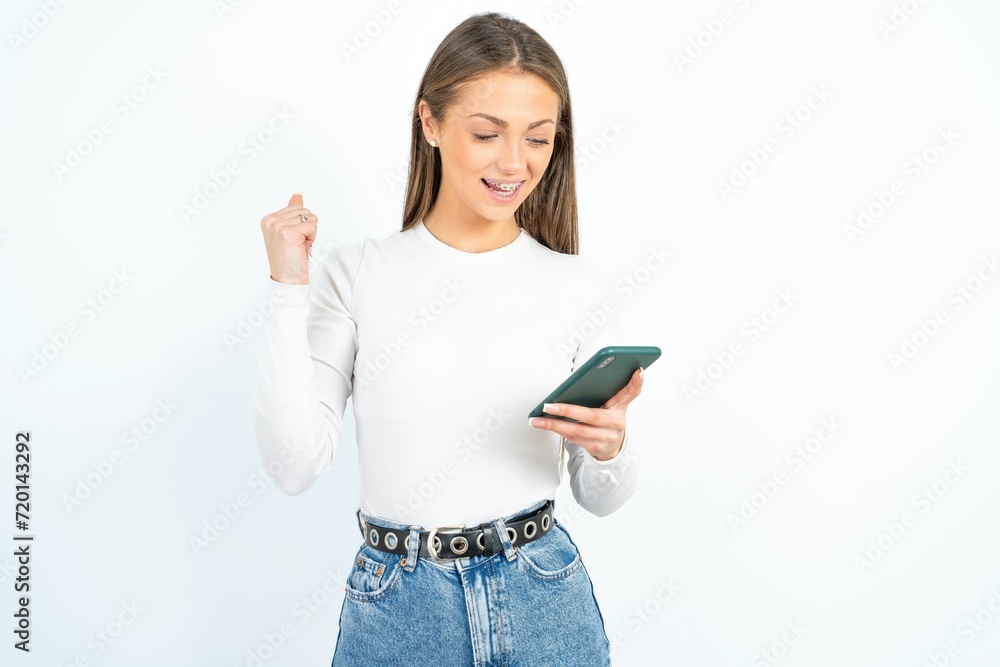 young beautiful girl wearing white T-shirt holding in hands cell and rising his fist up being excited after reading good news. young beautiful girl wearing white T-shirt holding in hands cell and rising his fist up being excited after reading good news.