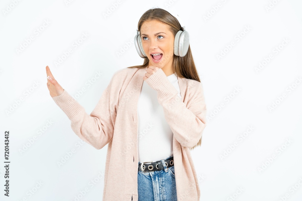 Happy beautiful girl wearing casual clothes sings favourite song keeps hand near mouth as if microphone wears wireless headphones, listens music Happy beautiful girl wearing casual clothes sings favourite song keeps hand near mouth as if microphone wears wireless headphones, listens music
