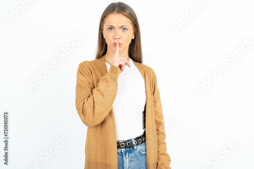 Surprised beautiful girl wearing casual clothes makes silence gesture, keeps finger over lips and looks mysterious at camera photo