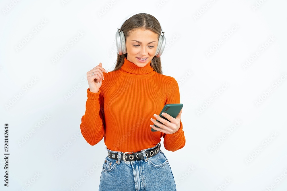 Happy young beautiful caucasian woman wearing orange turtleneck feels good while focused in screen of smartphone. People, technology, lifestyle