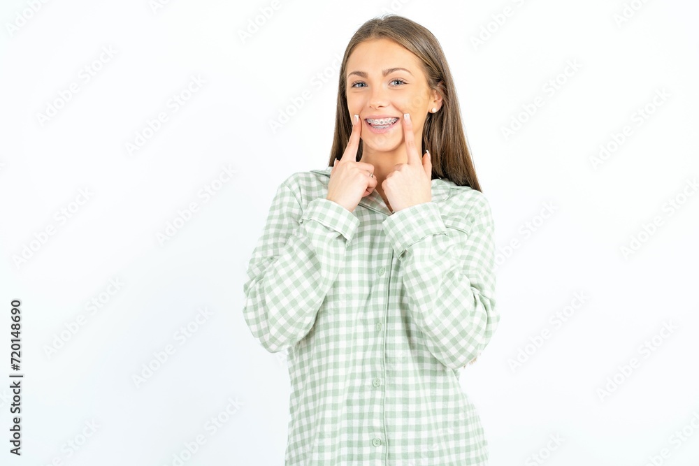 Happy Beautiful young woman wearing green plaid pyjama with toothy smile, keeps index fingers near mouth, fingers pointing and forcing cheerful smile Happy Beautiful young woman wearing green plaid pyjama with toothy smile, keeps index fingers near mouth, fingers pointing and forcing cheerful smile
