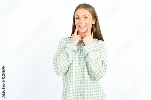 Happy Beautiful young woman wearing green plaid pyjama with toothy smile, keeps index fingers near mouth, fingers pointing and forcing cheerful smile