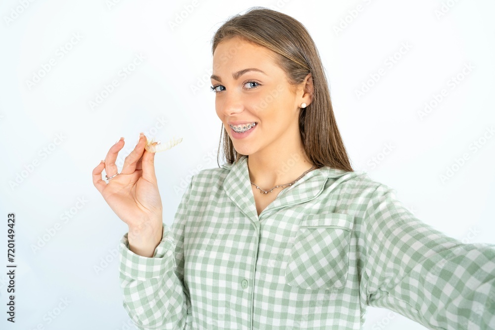 Beautiful young woman wearing green plaid pyjama make selfie holding an invisible braces aligner, recommending this new treatment. Dental healthcare concept. Beautiful young woman wearing green plaid pyjama make selfie holding an invisible braces aligner, recommending this new treatment. Dental healthcare concept.