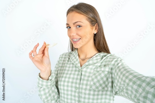 Beautiful young woman wearing green plaid pyjama make selfie holding an invisible braces aligner, recommending this new treatment. Dental healthcare concept.