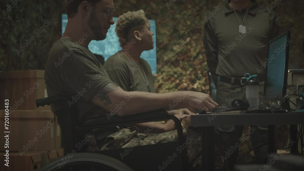 Tilt up lowkey shot of Caucasian male military worker in wheelchair greeting colleagues while approaching to work desk with monitoring program on computer, working in military control service office Tilt up lowkey shot of Caucasian male military worker in wheelchair greeting colleagues while approaching to work desk with monitoring program on computer, working in military control service office