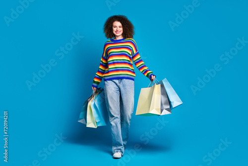 Full length photo of lovely young lady walk hold shopping bags wear trendy rainbow print garment isolated on blue color background