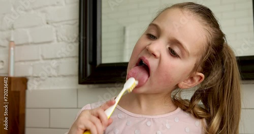 Portrait of a three-year-old girl with a ponytail in a pink sweater brushes her tongue with a yellow toothbrush in the morning at home in the bathroom