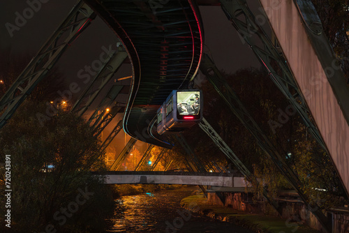 Obraz na płótnie Night cityscape of Wuppertal, North Rhine-Westphalia, Germany: Schwebebahn (the oldest in the world electric elevated suspension railway with hanging cars) with passeners is moving over the river. w salonie